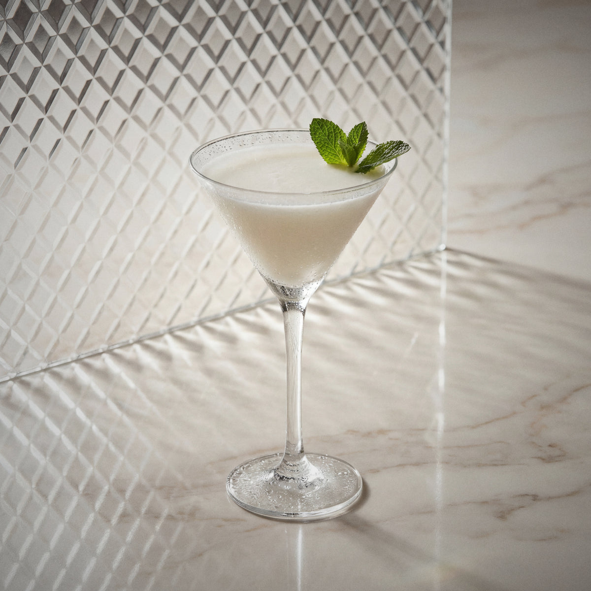 albino grasshopper cocktail with natural yoghurt liqueur, peppermint white liqueur, and cacao white liqueur in a martini glass with a mint leaf on a marble surface.