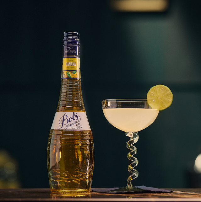 Bottle of Bols Banana Liqueur next to a cocktail with a lime wheel on a dark background