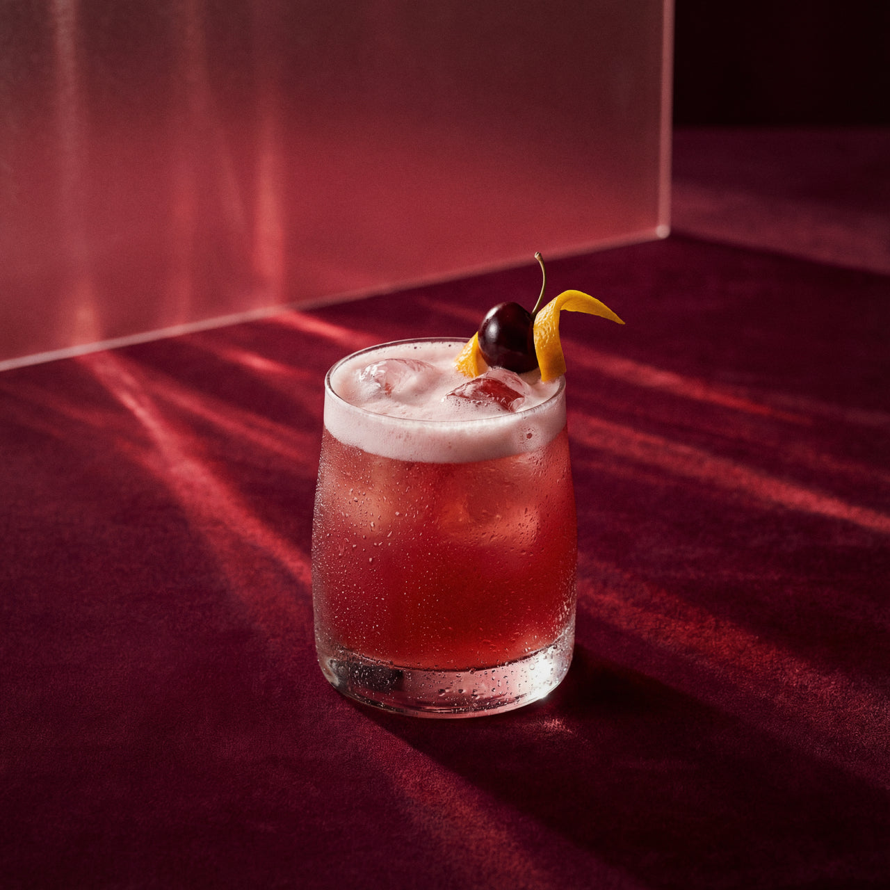 cherry sour cocktail with cherry brandy liqueur and a cherry and lemon peel on a dark red surface