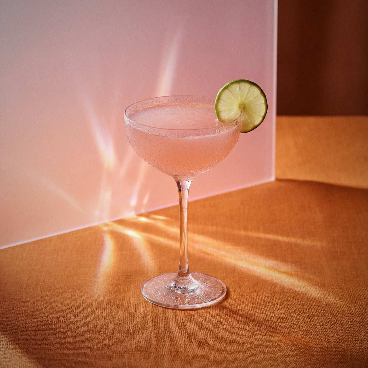 hemingway daiquiri cocktail with pink grapefruit liqueur and maraschino liqueur in a coupe glass and with a lime wheel on a textured pink surface