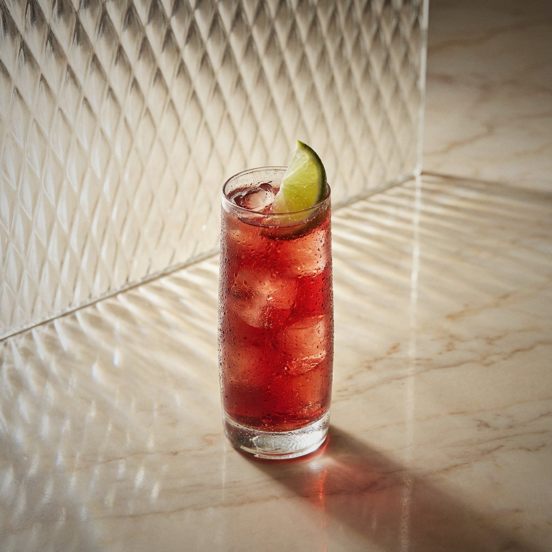 vanilla berry cooler cocktail with ice and a lime wedge and strawberry liqueur and vanilla liqueur in a highball glass on a marble surface.