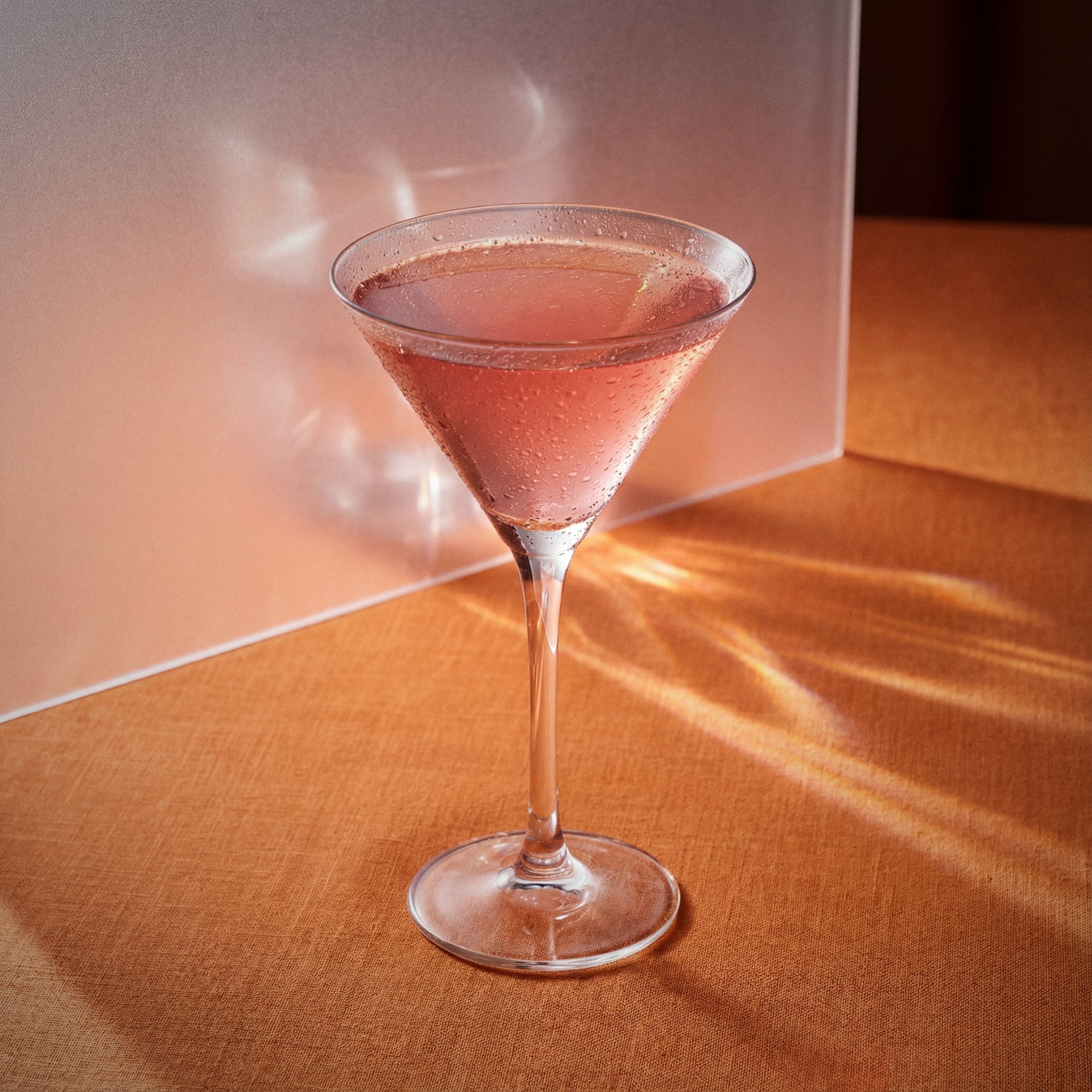 Verano cocktail in a martini glass with pink grapefruit liqueur on a wooden surface with sunlight casting shadows