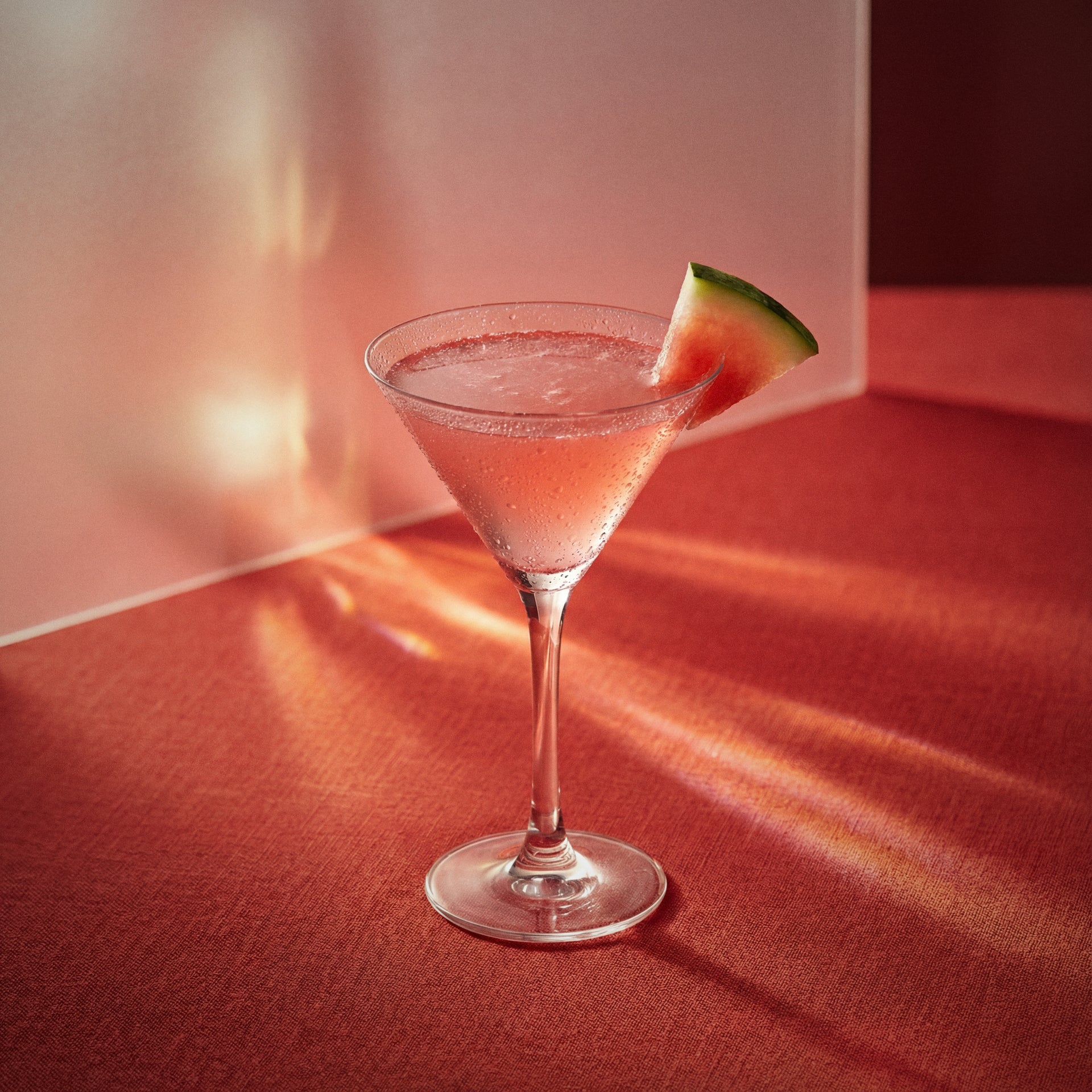 Watermelon Cosmopolitan cocktail with watermelon liqueur in a martini glass with a watermelon slice on a red surface
